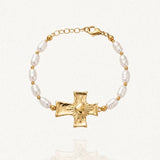 Bracelet doré composé de perles baroques blanches et de petites billes, centré sur une croix texturée avec un cœur rayonnant. Fermoir mousqueton et chaînette d'extension visibles.