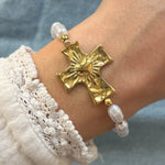 Bracelet de perles blanches irrégulières au poignet. Grande croix dorée martelée avec un cœur rayonnant. Détail manche dentelle.