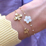 Deux bracelets en acier inoxydable sur un poignet: un doré à fleurs, l'autre doré à perles blanches et fleur nacre.