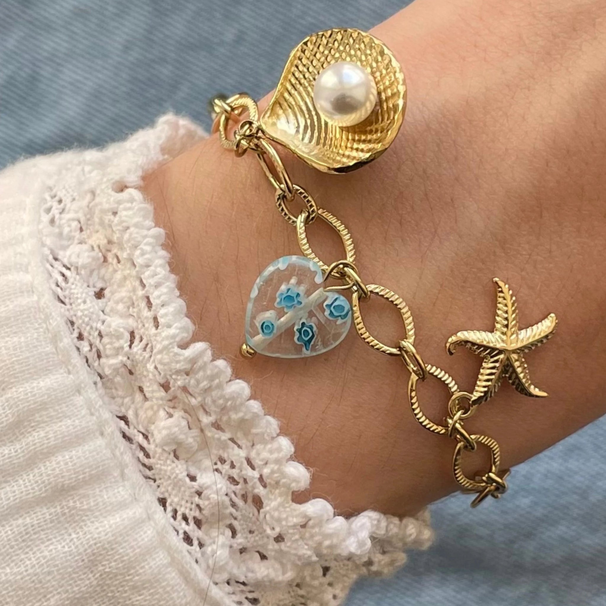 Gros plan bracelet chaîne en acier inoxydable doré orné de breloques marines : coquillage avec perle, cœur bleu translucide et étoile de mer, porté au poignet.