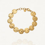 Bracelet DANIELLE en acier inoxydable doré. Il se compose de douze médaillons ronds et martelés, reliés en chaîne. Le bracelet possède un fermoir mousqueton et une chaînette d'extension. Il est posé sur un fond blanc.