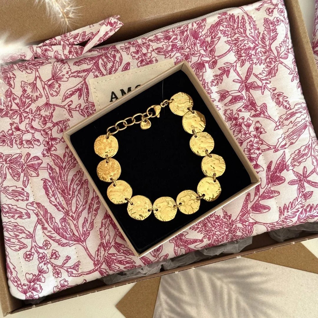 Bracelet doré à maillons disques martelés dans écrin noir, posé sur une pochette matelassée motif toile de Jouy rose.