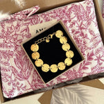 Bracelet doré à maillons disques martelés dans écrin noir, posé sur une pochette matelassée motif toile de Jouy rose.