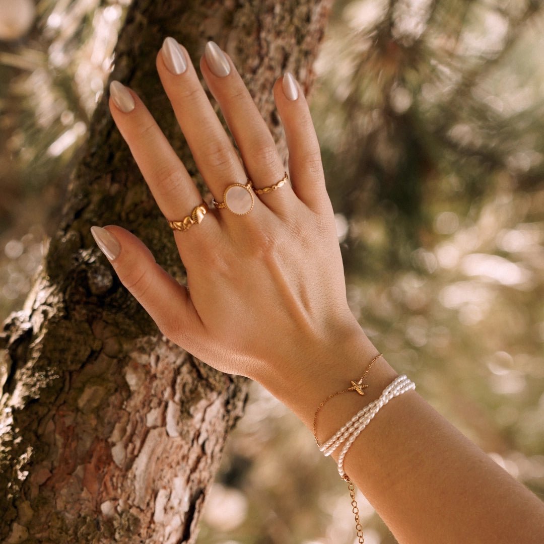 Bracelet femme en acier inoxydable doré avec perles blanches et pendentif étoile de mer, main sur tronc d'arbre.