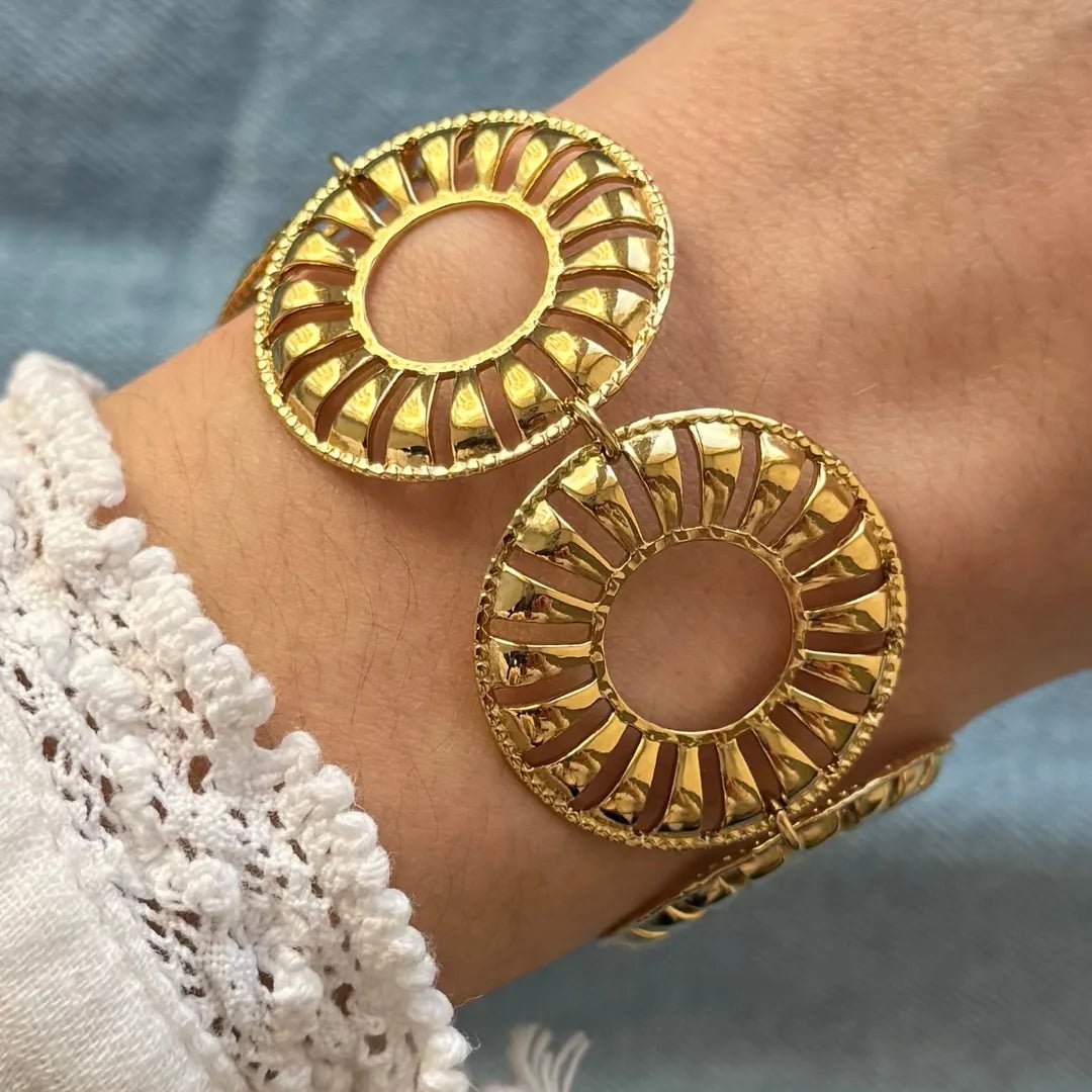 Bracelet doré à maillons circulaires ajourés, motif rayons striés et bordure perlée, sur un poignet, manche dentelle blanche.