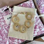 Bracelet doré à 5 maillons ronds ajourés motif soleil, posé sur pochette beige dans un emballage cadeau floral rose et marron.