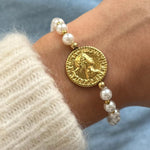 Gros plan bracelet perles blanches et petites billes dorées avec médaillon central pièce de monnaie antique porté au poignet.