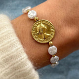 Gros plan bracelet perles blanches d'eau douce et billes dorées avec médaillon central pièce de monnaie antique porté au poignet.