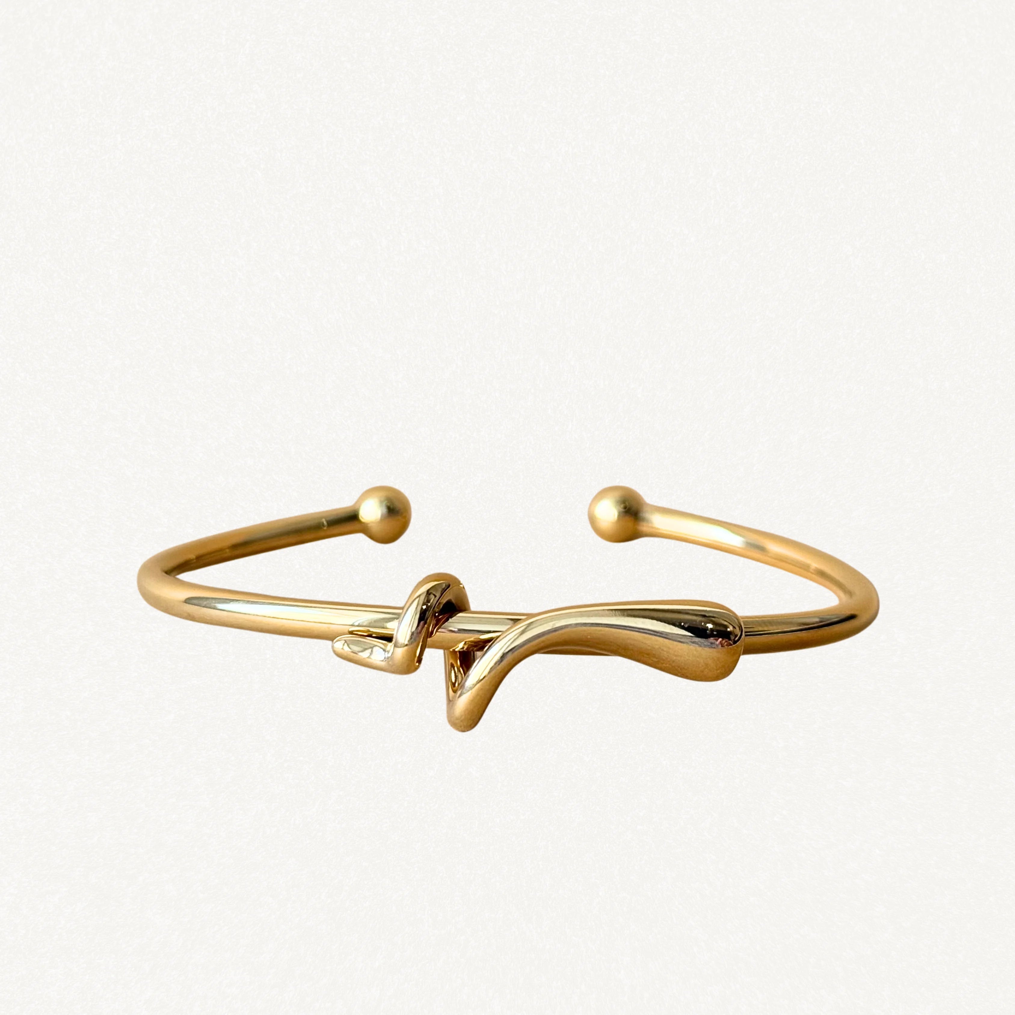 Bracelet jonc rigide ouvert en métal doré avec motif nœud central asymétrique et extrémités boules sur fond blanc.