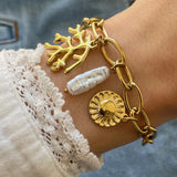 Gros plan bracelet grosse maille en acier inoxydable doré avec breloques branche de corail, perle baroque allongée et médaille soleil porté au poignet.