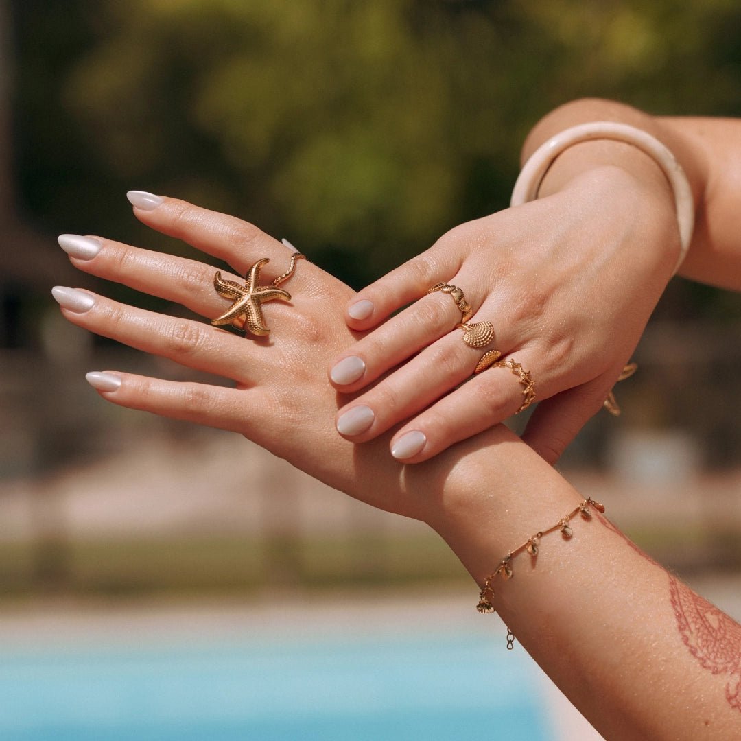 Bracelet fin en acier inoxydable doré avec de petits pendentifs, porté sur un poignet féminin au bord d'une piscine.