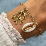 Gros plan bracelet chaîne maillons allongés en acier inoxydable doré avec breloques branche de corail et coquillage cauri naturel porté au poignet.