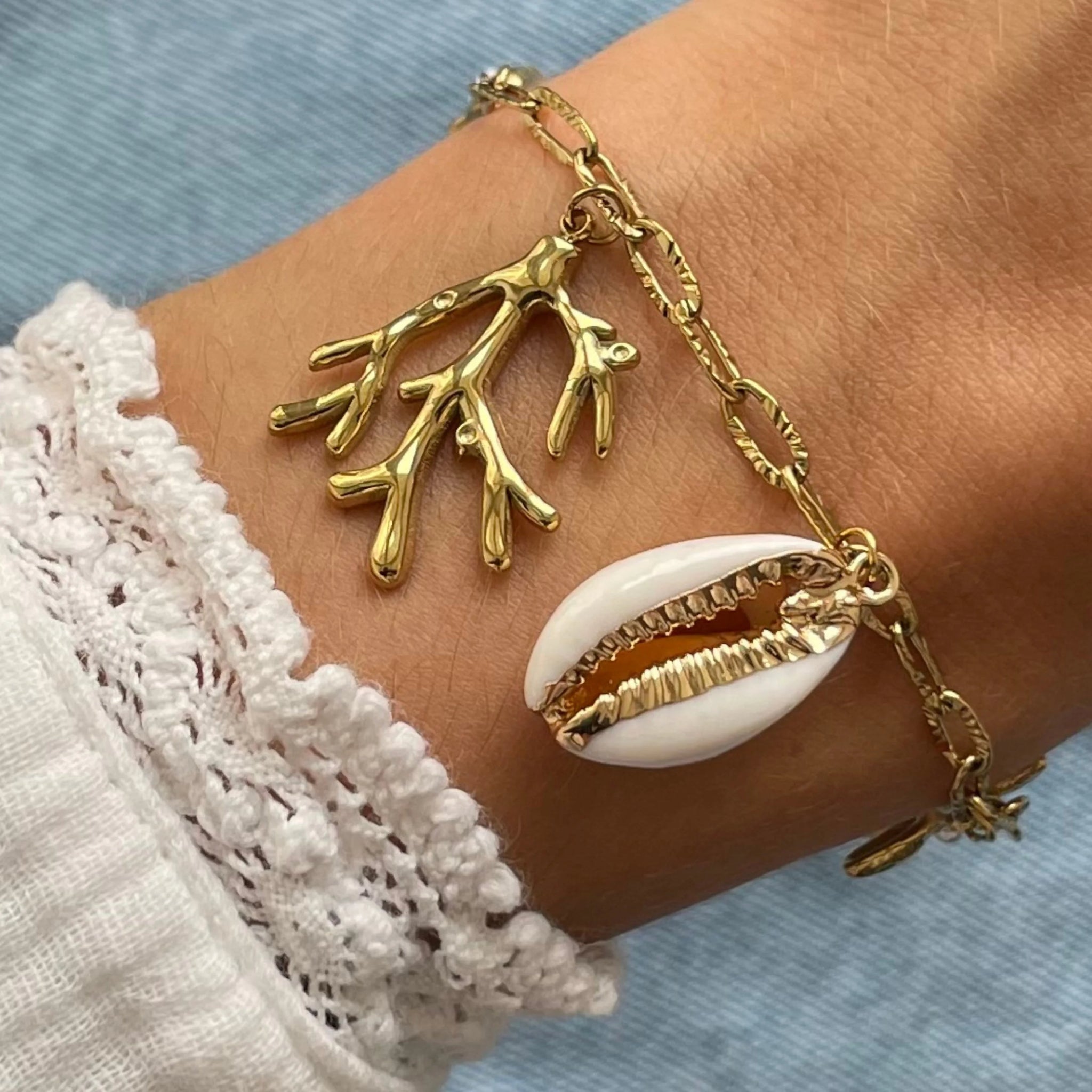 Gros plan bracelet chaîne maillons allongés en acier inoxydable doré avec breloques branche de corail et coquillage cauri naturel porté au poignet.