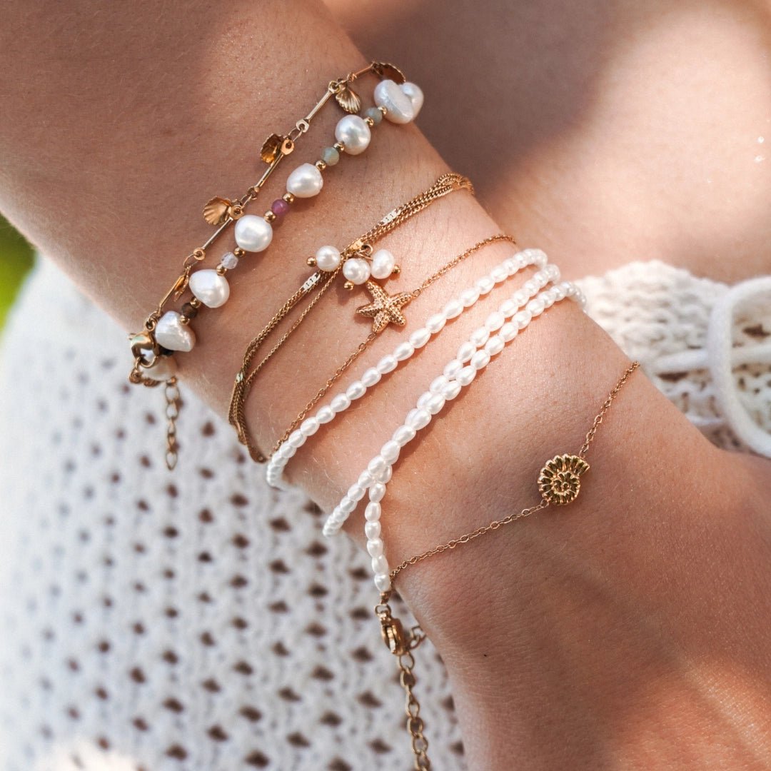 Ensemble de bracelets en acier inoxydable doré, perles, coquillages et étoile de mer sur poignet de femme.