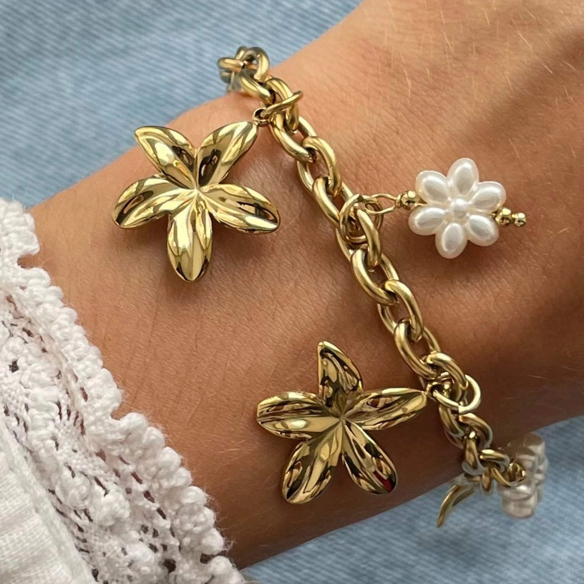 Gros plan bracelet chaîne maillons épais en acier inoxydable doré avec breloques grandes fleurs dorées et petites fleurs en perles blanches porté au poignet.