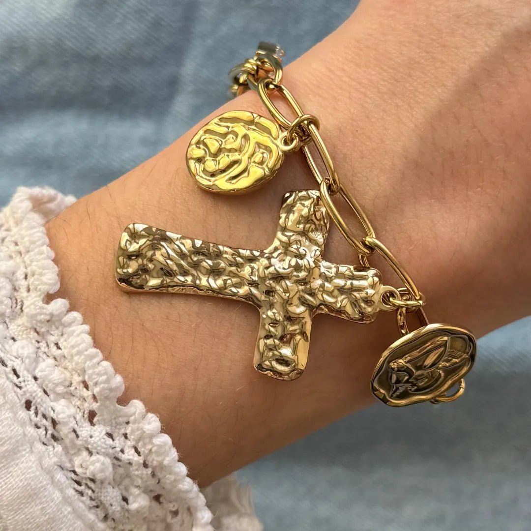 Bracelet à maillons dorés sur poignet, avec grande croix martelée, charme rond motif rose et charme mains priantes.