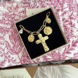Bracelet doré à chaîne avec grande croix texturée, médaillons angelots et cristal goutte, dans une boîte sur fond floral rose.
