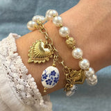 Bracelet avec perles blanches, chaîne dorée, cœur sacré doré, et pendentif ovale en céramique blanche à fleurs bleues, sur un poignet.