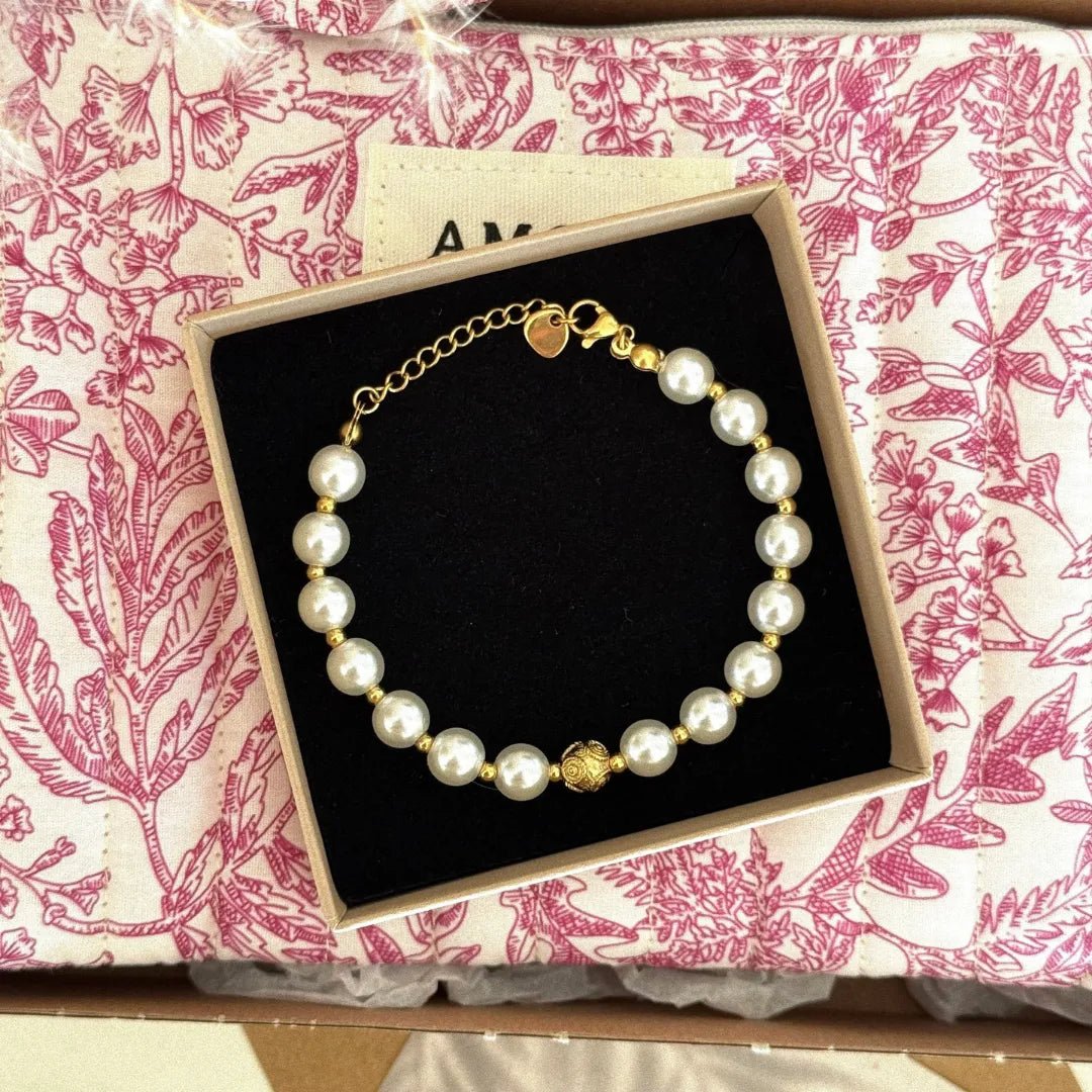 Bracelet de perles blanches et petites billes dorées avec fermoir or, breloque ouvragée, dans boîte sur fond toile de Jouy rose.