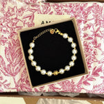 Bracelet de perles blanches et petites billes dorées avec fermoir or, breloque ouvragée, dans boîte sur fond toile de Jouy rose.