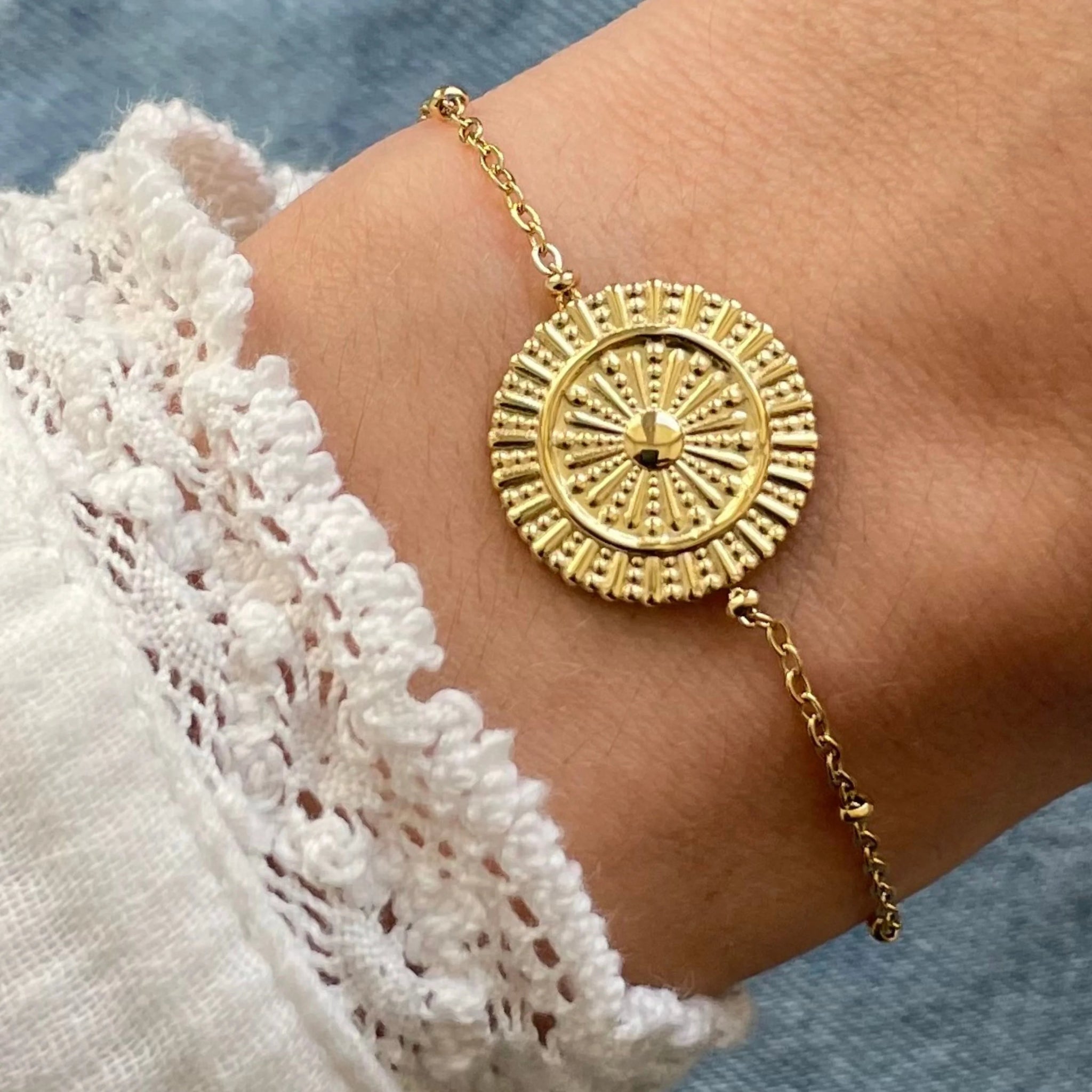Gros plan bracelet chaîne fine en acier inoxydable doré avec grand médaillon rond motif soleil ou roue ethnique porté au poignet.