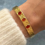 Gros plan bracelet jonc ouvert en acier inoxydable doré effet martelé orné de trois pierres rouges ovales porté au poignet.
