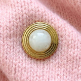 Détail de la broche ronde Angela finition dorée et résine nacrée blanche, portée sur un pull en maille rose.