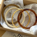 Coffret cadeau ouvert présentant un assortiment de bracelets joncs en résine et doré disposés délicatement dans du papier de soie blanc.