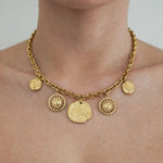 Collier chaîne maillons épaisse dorée, 5 charms pendants: 2 soleils ronds, 2 pièces antiques à profil, 1 grand médaillon avec symboles gravés.