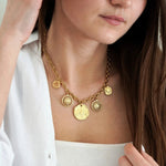 Collier chaîne épaisse dorée avec 6 charms: 2 soleils stylisés et 4 médaillons, dont une pièce antique, sur une femme.