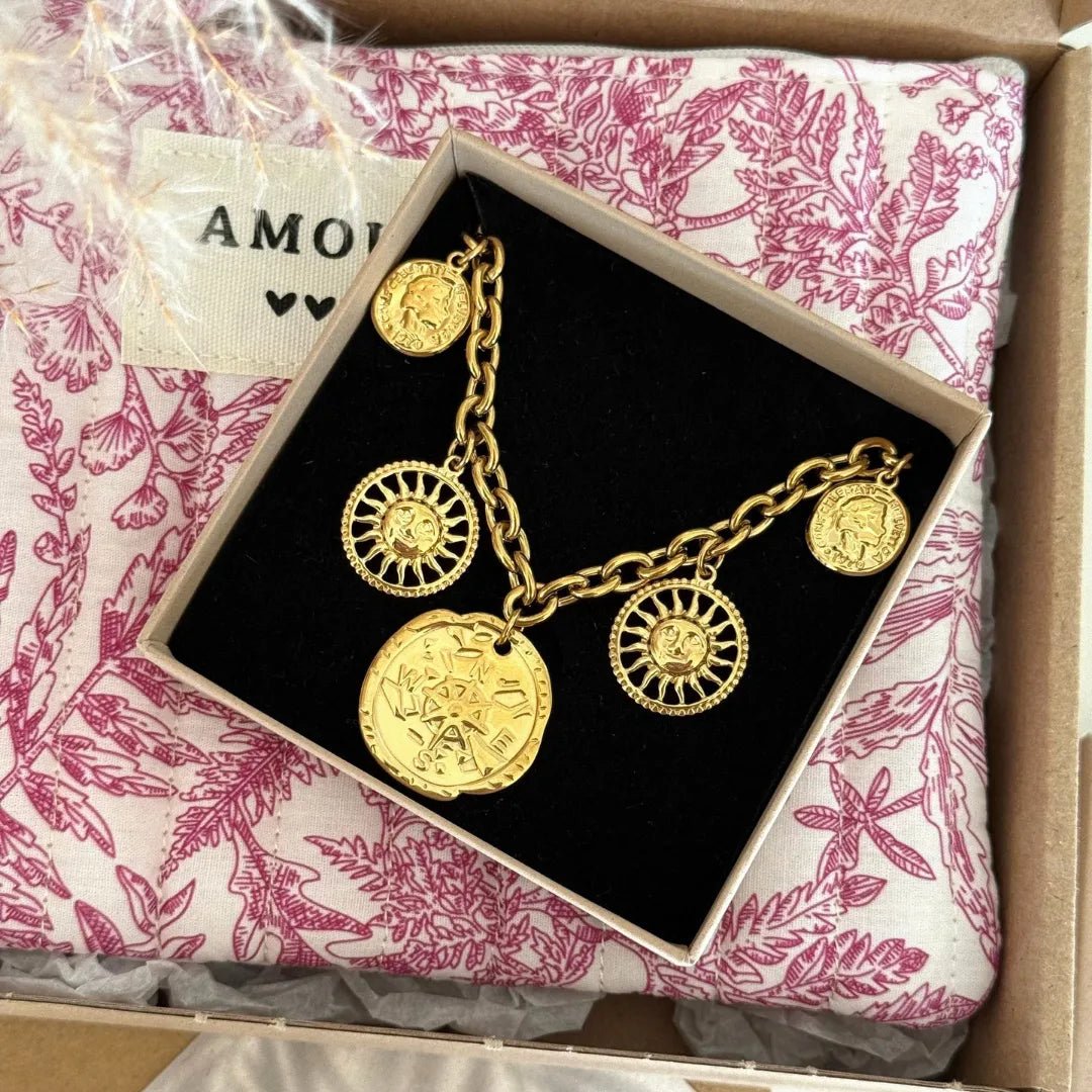 Bracelet chaîne dorée avec 2 charmes soleils, 2 médailles visages, 1 rose des vents, dans sa boîte noire sur tissu toile de Jouy.