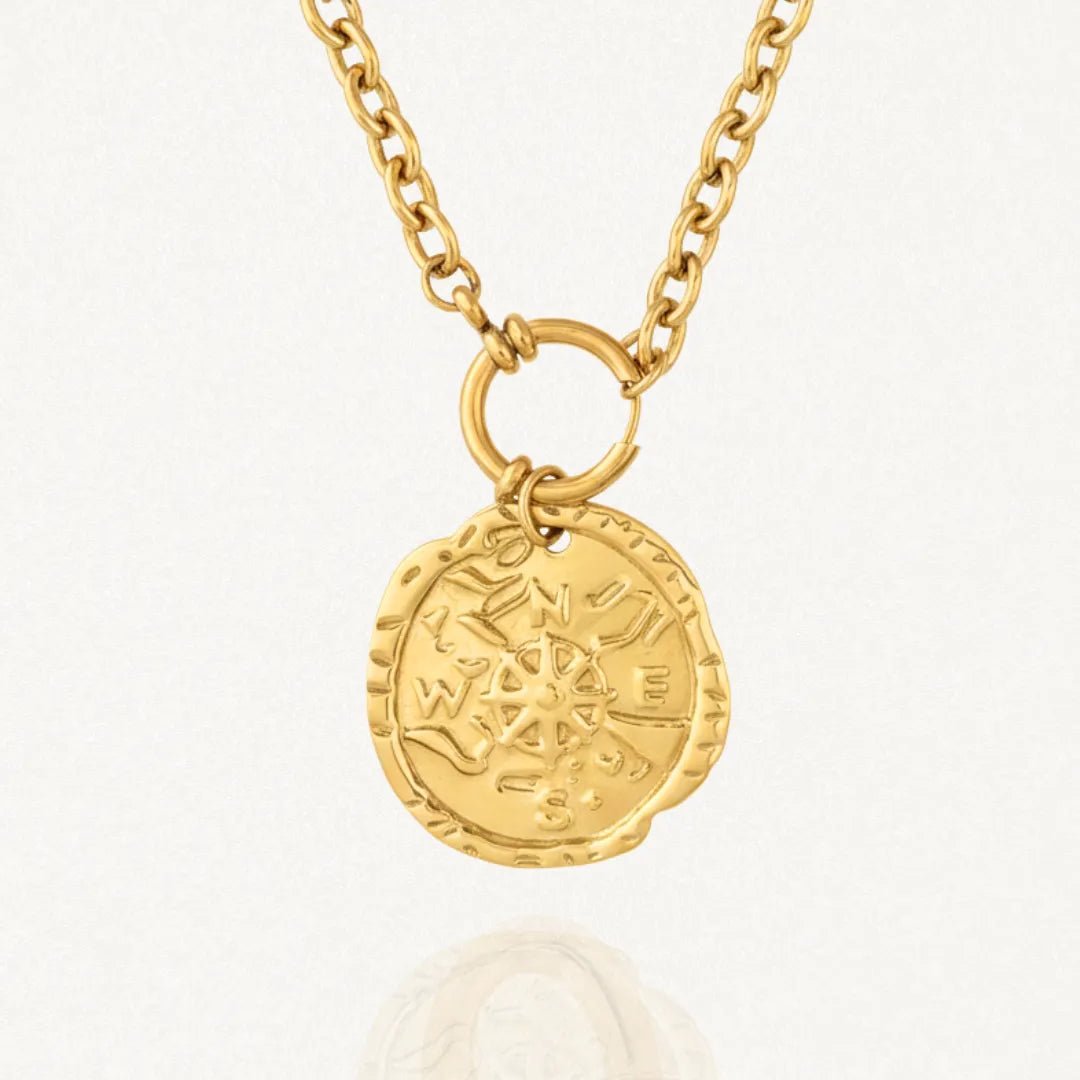 Collier doré en acier inoxydable avec une chaîne à maillons et un pendentif médaille ronde texturée. La médaille est gravée d'une boussole et des points cardinaux N, E, S, W.