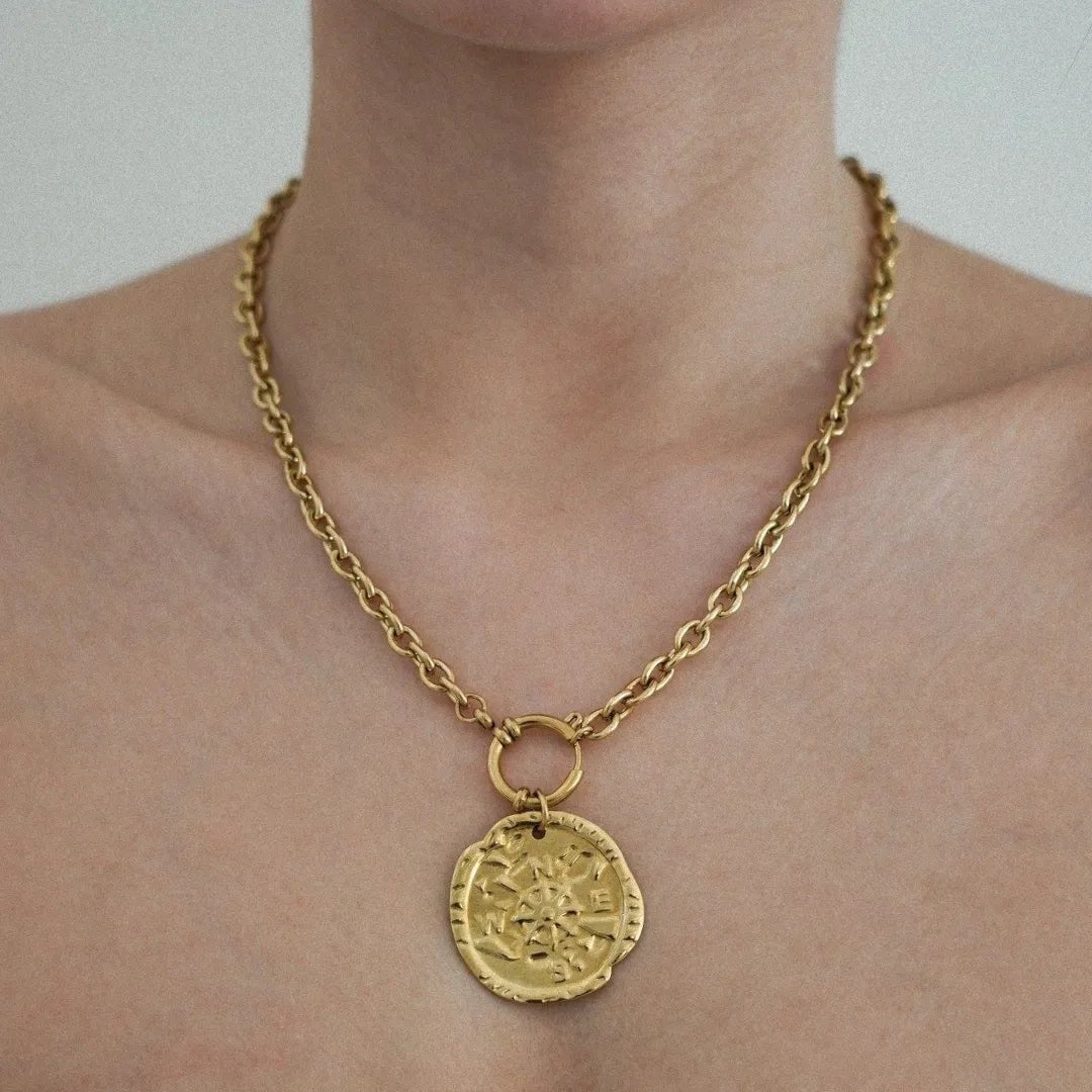 Collier chaîne dorée épaisse à maillons ovales, pendentif médaille irrégulière rose des vents, porté sur cou de femme.
