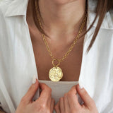 Collier chaîne or épaisse avec pendentif médaille d'or texturée, porté par femme en chemise blanche, mains au col.