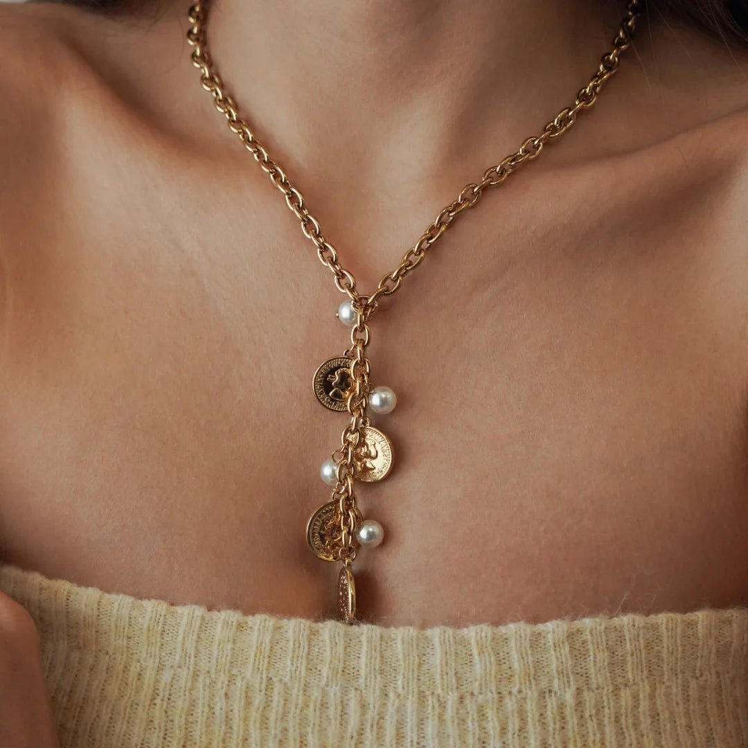 Collier cravate plongeant forme Y en acier inoxydable doré orné de médailles pièces de monnaie et de perles blanches porté au cou.