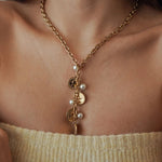 Collier cravate plongeant forme Y en acier inoxydable doré orné de médailles pièces de monnaie et de perles blanches porté au cou.