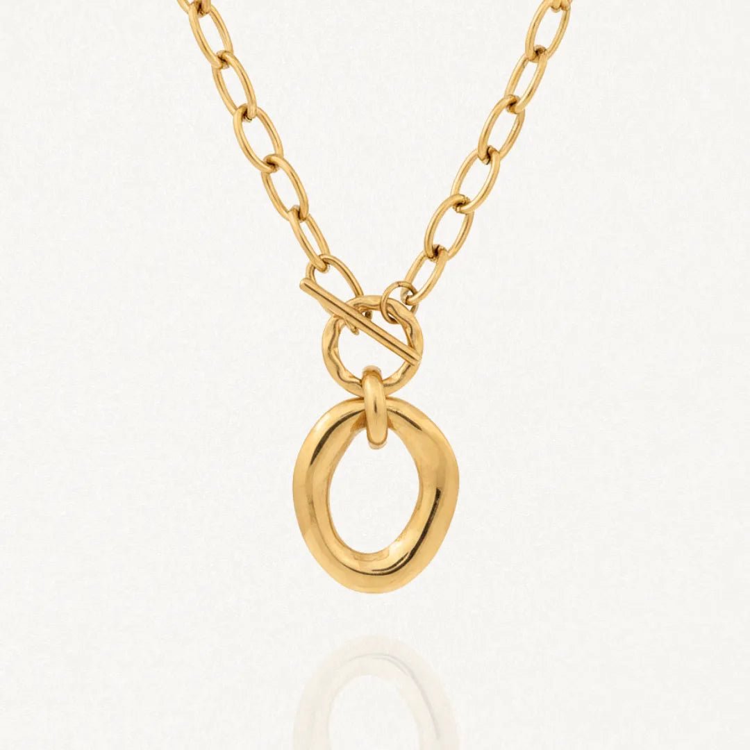 Collier BENEDICTE en acier inoxydable doré. Chaîne à maillons ovales, fermoir toggle et grand pendentif ovale de forme organique irrégulière. Posé sur fond blanc.