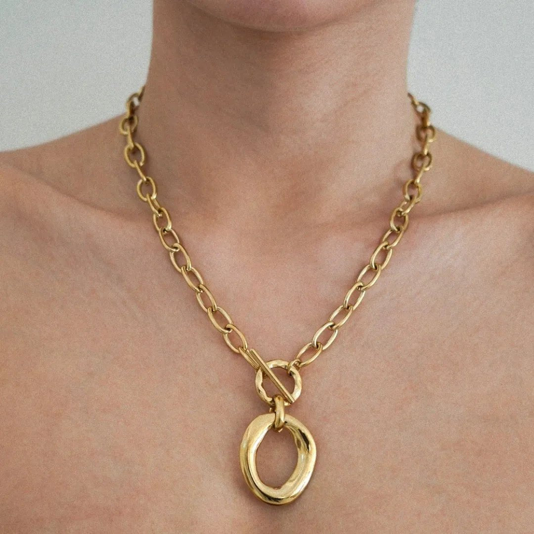 Collier chaîne dorée épaisse à maillons ovales, fermoir toggle et pendentif anneau doré organique sur un cou.
