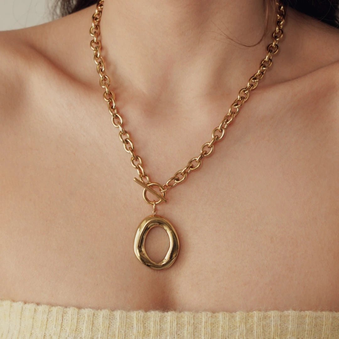Collier chaîne dorée épaisse à maillons ovales, fermoir toggle et pendentif anneau doré organique sur un cou.