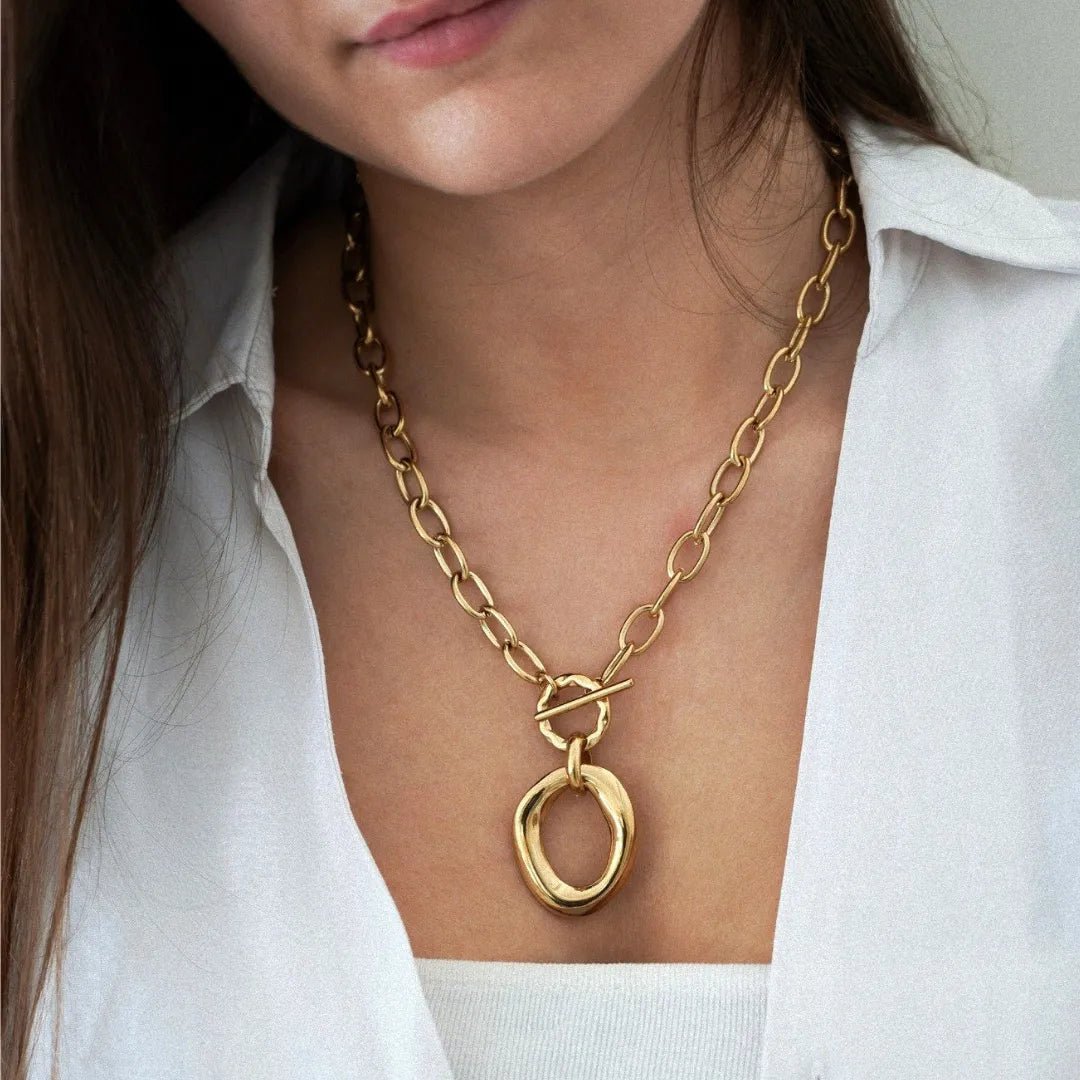 Collier chaîne dorée à maillons ovales, fermoir toggle et pendentif anneau ovale irrégulier, porté sur décolleté de femme.