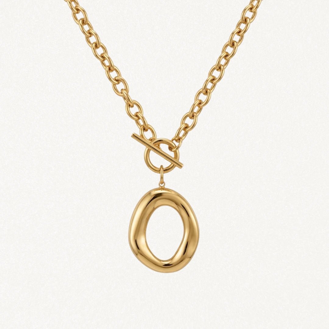 Collier BENEDICTE en acier inoxydable doré. Chaîne à maillons ovales, fermoir toggle et grand pendentif ovale de forme organique irrégulière. Posé sur fond blanc.