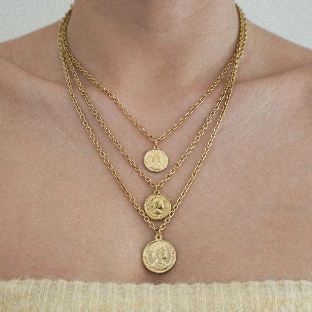 Trois colliers chaînes dorées superposés avec pendentifs médaillons pièces de monnaie, portés sur un buste féminin.