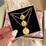 Collier doré trois chaînes superposées, trois médaillons profil reine. Tenu en boîte par une main manucurée blanche.