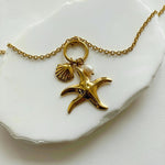 Gros plan sur les breloques du collier Bruna : étoile de mer dorée, petit coquillage et perle naturelle posés sur une pierre blanche.