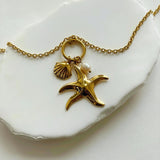 Gros plan sur les breloques du collier Bruna : étoile de mer dorée, petit coquillage et perle naturelle posés sur une pierre blanche.