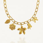 Gros plan sur les breloques du collier : fleurs, nœud papillon et feuille sur chaîne dorée fond blanc.