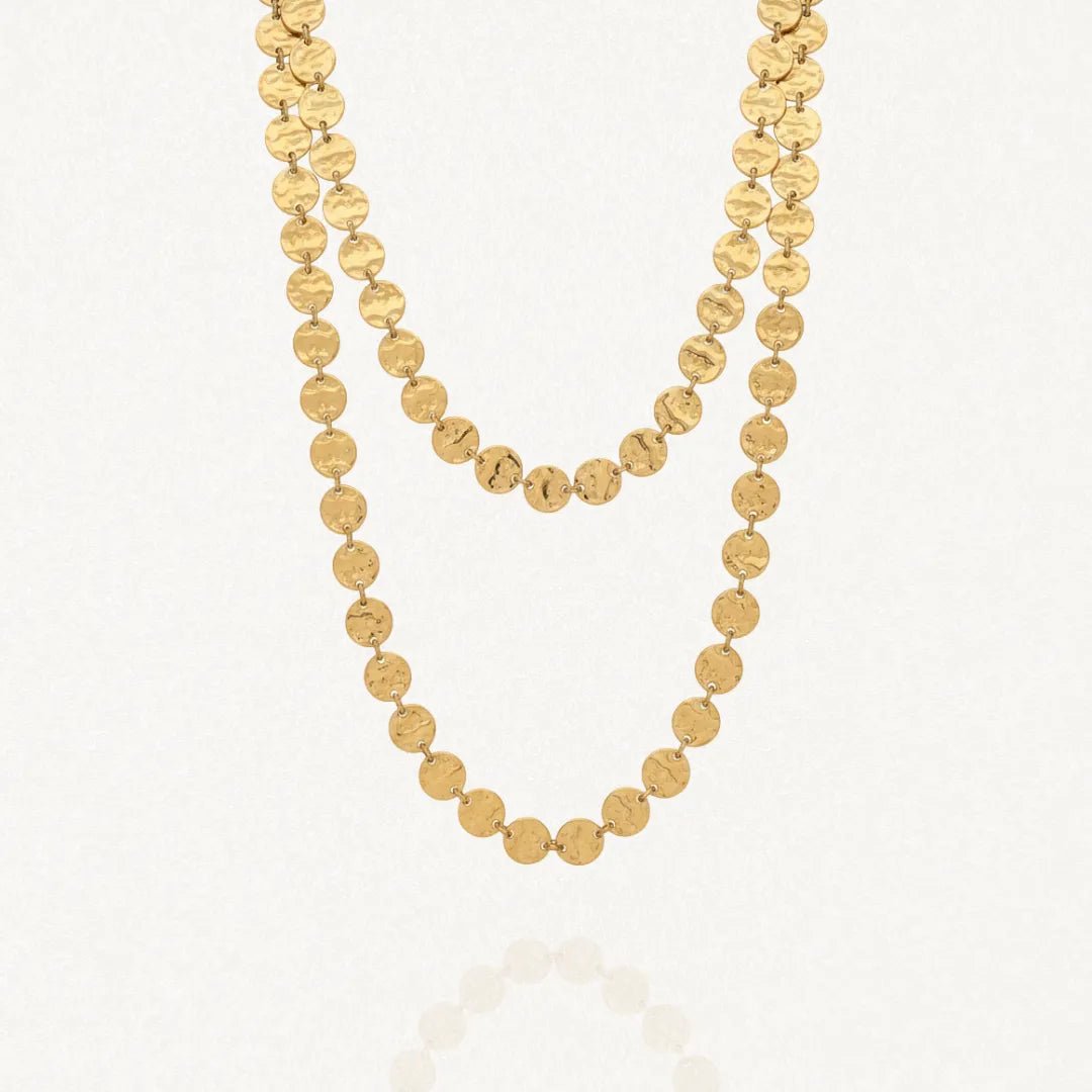 Collier long doré à deux rangs composé de multiples disques ronds martelés en acier inoxydable, posé sur un fond blanc avec un léger reflet.