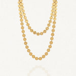 Collier long doré à deux rangs composé de multiples disques ronds martelés en acier inoxydable, posé sur un fond blanc avec un léger reflet.