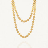 Collier long doré à deux rangs composé de multiples disques ronds martelés en acier inoxydable, posé sur un fond blanc avec un léger reflet.