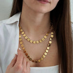 Collier double doré à longs maillons de disques ronds martelés, porté par une femme en chemise blanche et débardeur.